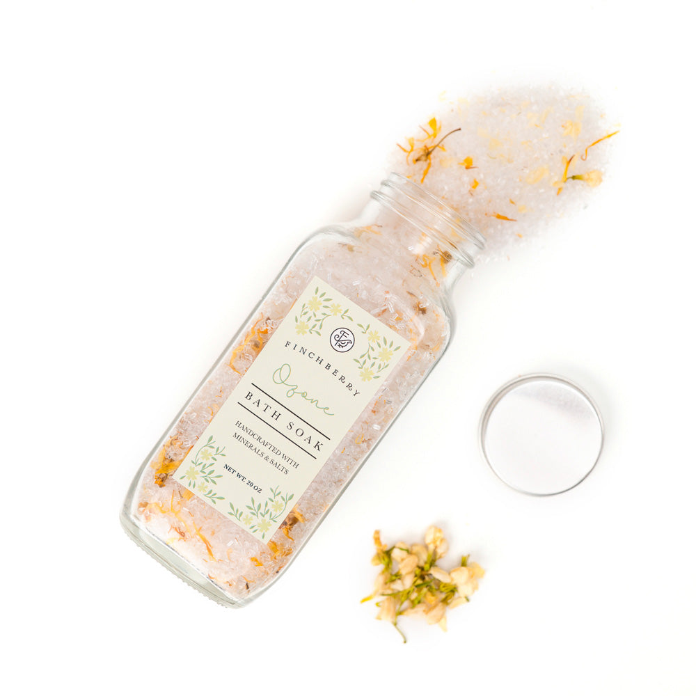 Finchberry Ozone Bath Soak shown pouring out of the glass apothecary style bottle. The bath salts have herbs and flowers without.